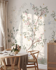 May include: A dining room scene featuring a floral wallpaper with a tree design, pink roses, and yellow fruit. The room includes a round table set for dining, wooden chairs, and a cabinet. Soft, natural light filters through the window.
